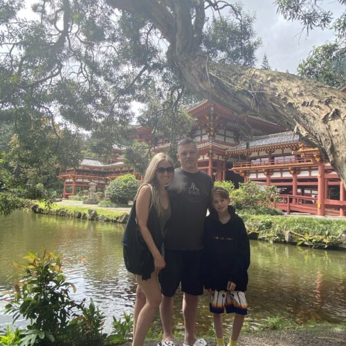 A blond woman with a black purse, a man in a green tee shirt and black shorts and a boy in a black sweatshirt and shorts under a tree with a red temple in the background